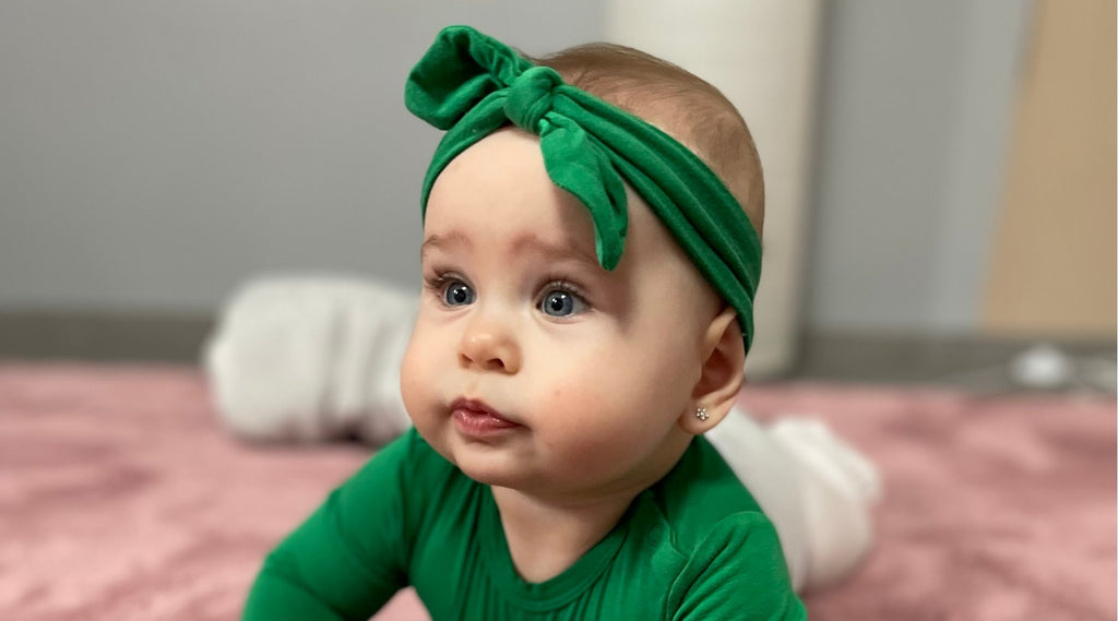 Kislány baba zöld bodyban és zöld masniban hasal a rózsaszín szőnyegen