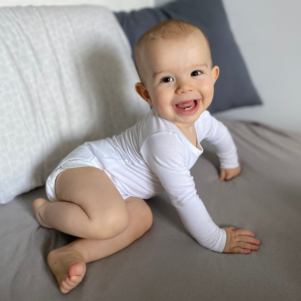 Kisbaba fehérbodyban mosolyogva ül az ágyon