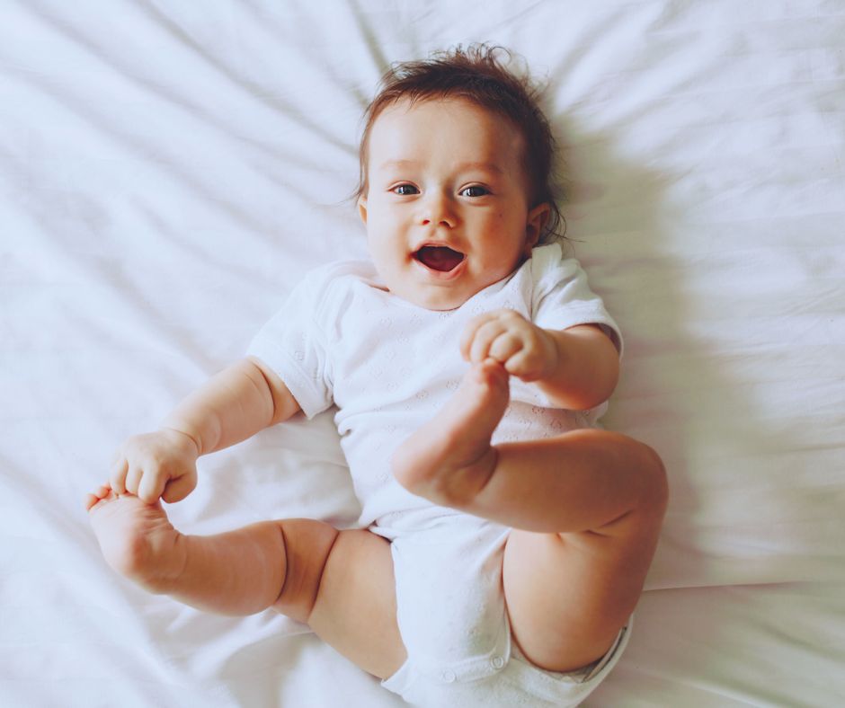 Mozgékony kisbaba fekszik a hátán, kezeivel lábacskáit fogja és mosolyog