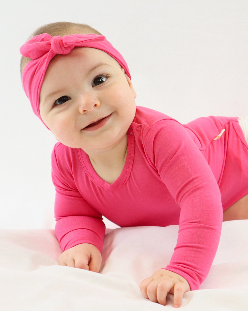 Kicsi baba oldalt fekszik épp felkönyökölve ránk mosolyog rózsaszín body-t és masni fejpántot visel a LiaaBébé-től