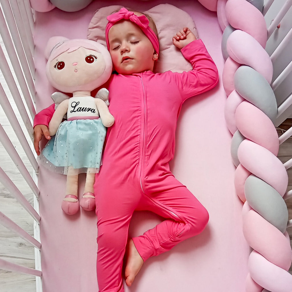 Kislány rózsaszn egyberészes bambusz pizsamában alszik a rózsaszín kiságyában a babájával