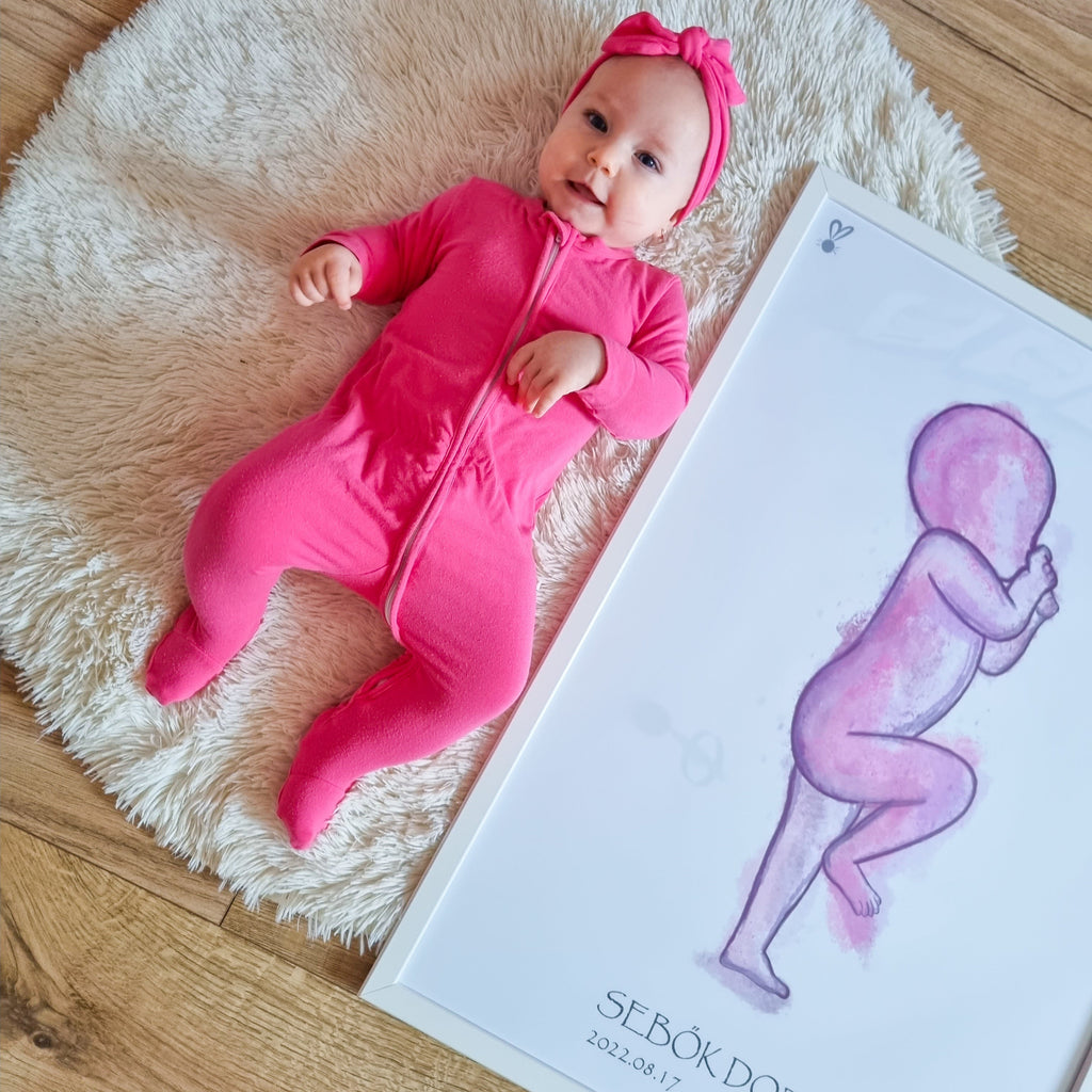 Rózsaszn rugdalózóvan egy kicsi baba fekszik egy bekeretezett születési méret arányos babaposzter mellett